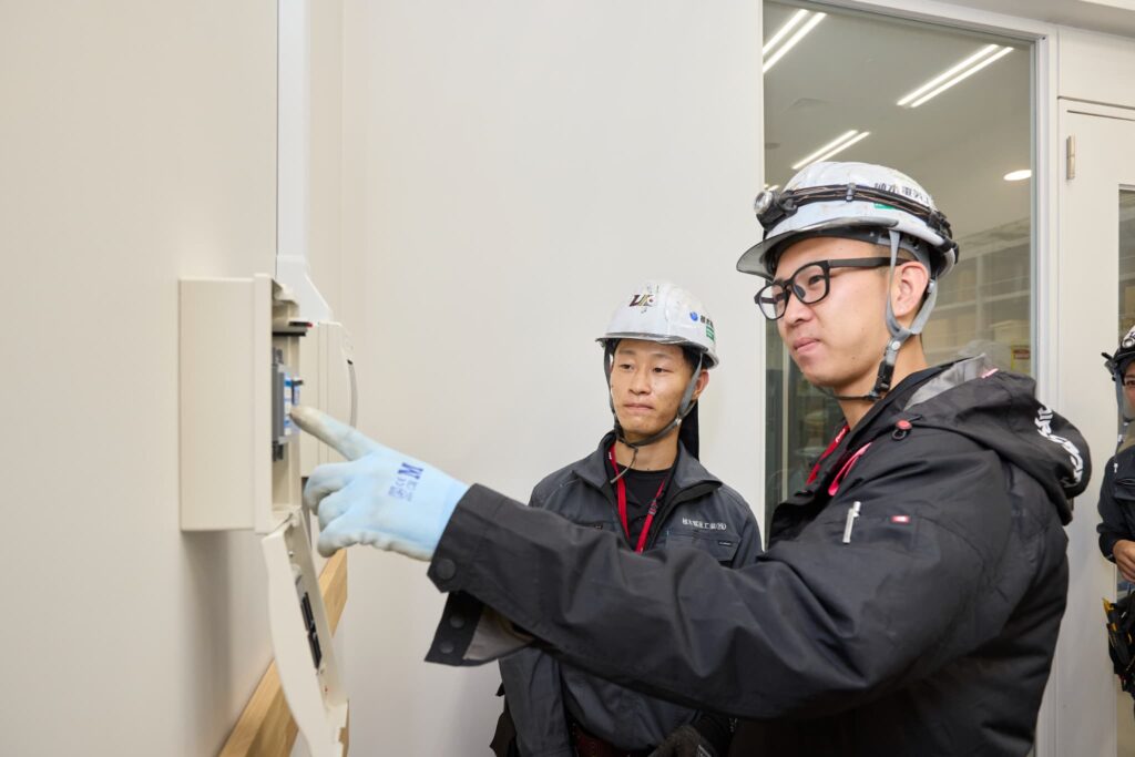 電気工事士が分電盤を指しながら指導している現場